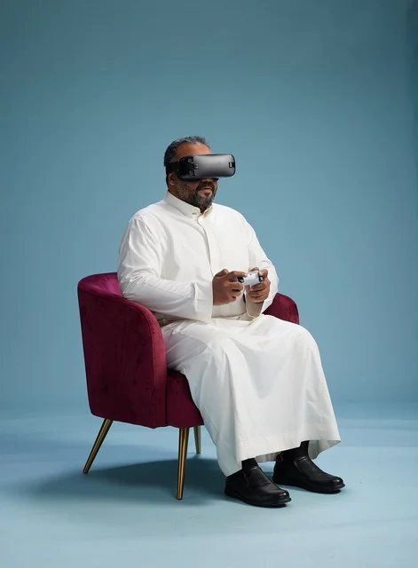 A portrait of a Saudi man wearing virtual reality glasses and holding a controller, enjoying video games, with a blue background.
