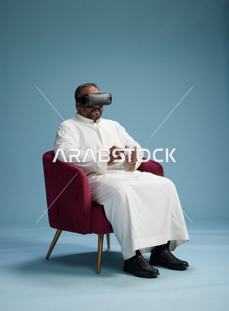 بورتريه لرجل سعودي يرتدي نظارة واقع افتراضي ويمسك بوحدة تحكم, الاستمتاع بالعاب الفيديو, خلفيه زرقاء