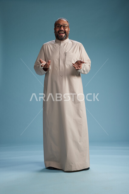 بورتريه لرجل عربي سعودي خليجي مبتسم يرتدي ثوب بيج يمسك بيده سبحه وينظر الى الكاميرا, كامل الجسم, خلفيه زرقاء