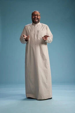 بورتريه لرجل عربي سعودي خليجي مبتسم يرتدي ثوب بيج يمسك بيده سبحه وينظر الى الكاميرا, كامل الجسم, خلفيه زرقاء