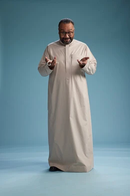 A portrait of a smiling Arab Saudi Gulf man wearing a beige thobe, holding a rosary in his hand and looking at the camera, full body, with a blue background.