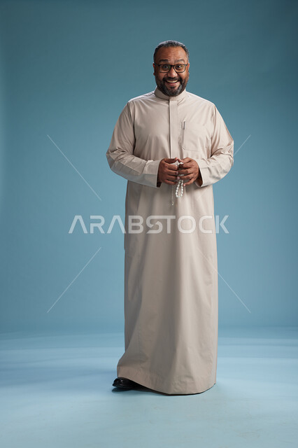 بورتريه لرجل عربي سعودي خليجي مبتسم يرتدي ثوب بيج يمسك بيده سبحه وينظر الى الكاميرا, كامل الجسم, خلفيه زرقاء
