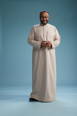 A portrait of a smiling Arab Saudi Gulf man wearing a beige thobe, holding a rosary in his hand and looking at the camera, full body, with a blue background.