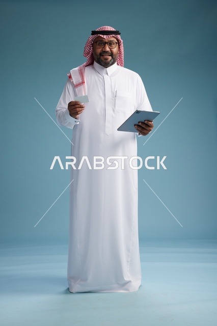 رجل سعودي بالزي التقليدي يستخدم جهاز لوحي وبطاقة ائتمان, مفهوم الدفع الالكتروني, الشراء عبر الونلاين, خلفيه زرقاء, كامل الجسم