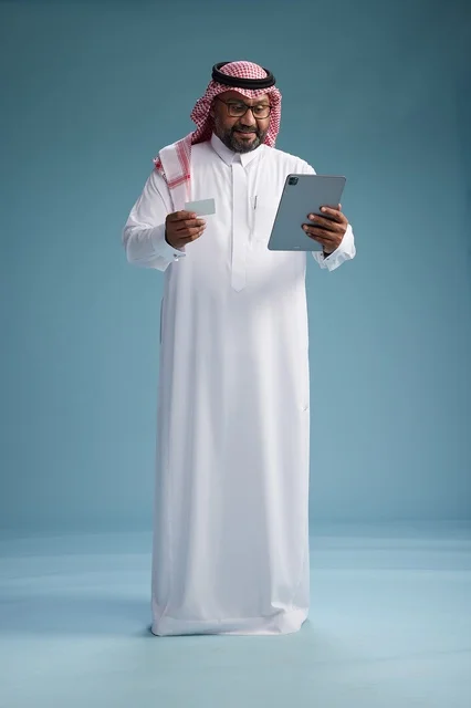 A Saudi man in traditional attire using a tablet and a credit card, the concept of electronic payment, online shopping, blue background, full body.