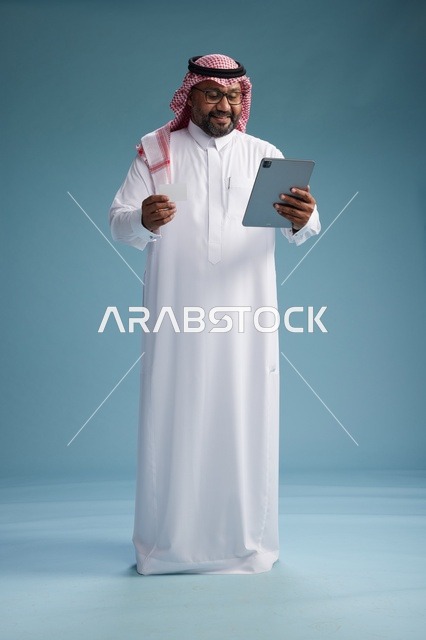 رجل سعودي بالزي التقليدي يستخدم جهاز لوحي وبطاقة ائتمان, مفهوم الدفع الالكتروني, الشراء عبر الونلاين, خلفيه زرقاء, كامل الجسم