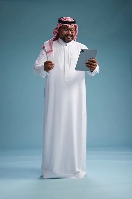 A Saudi man in traditional attire using a tablet and a credit card, the concept of electronic payment, online shopping, blue background, full body.