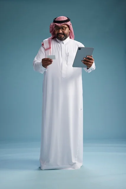 رجل سعودي بالزي التقليدي يستخدم جهاز لوحي وبطاقة ائتمان, مفهوم الدفع الالكتروني, الشراء عبر الونلاين, خلفيه زرقاء, كامل الجسم