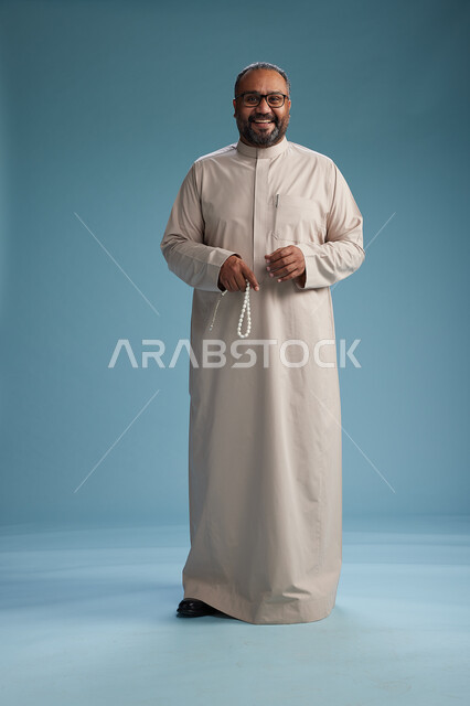 بورتريه لرجل عربي سعودي خليجي مبتسم يرتدي ثوب بيج يمسك بيده سبحه وينظر الى الكاميرا, كامل الجسم, خلفيه زرقاء