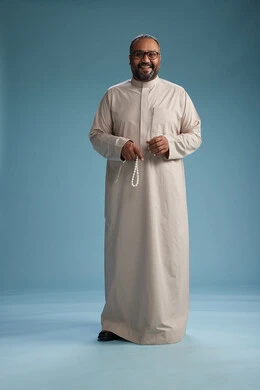 A portrait of a smiling Arab Saudi Gulf man wearing a beige thobe, holding a rosary in his hand and looking at the camera, full body, with a blue background.