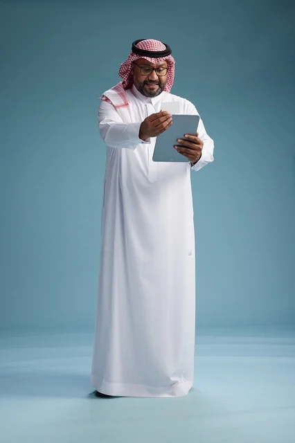 رجل سعودي بالزي التقليدي يستخدم جهاز لوحي وبطاقة ائتمان, مفهوم الدفع الالكتروني, الشراء عبر الونلاين, خلفيه زرقاء, كامل الجسم