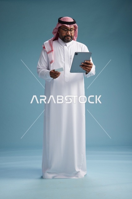 رجل سعودي بالزي التقليدي يستخدم جهاز لوحي وبطاقة ائتمان, مفهوم الدفع الالكتروني, الشراء عبر الونلاين, خلفيه زرقاء, كامل الجسم
