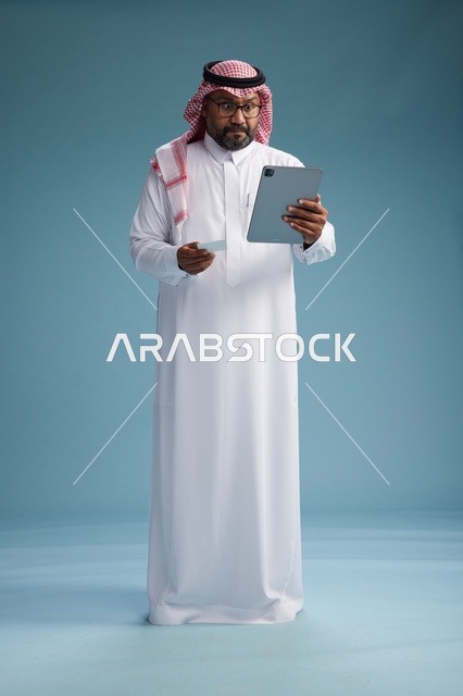 رجل سعودي بالزي التقليدي يستخدم جهاز لوحي وبطاقة ائتمان, مفهوم الدفع الالكتروني, الشراء عبر الونلاين, خلفيه زرقاء, كامل الجسم
