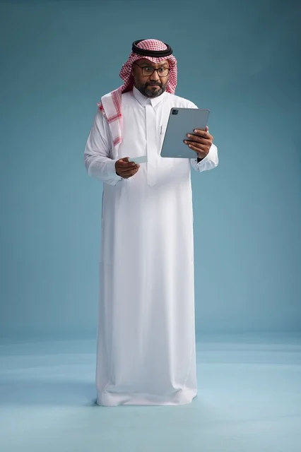 A Saudi man in traditional attire using a tablet and a credit card, the concept of electronic payment, online shopping, blue background, full body.