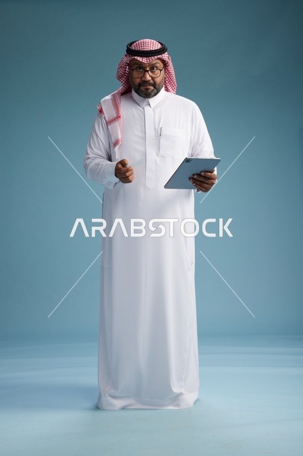 رجل سعودي بالزي التقليدي يستخدم جهاز لوحي وبطاقة ائتمان, مفهوم الدفع الالكتروني, الشراء عبر الونلاين, خلفيه زرقاء, كامل الجسم