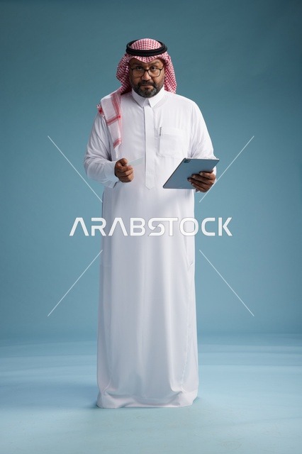 رجل سعودي بالزي التقليدي يستخدم جهاز لوحي وبطاقة ائتمان, مفهوم الدفع الالكتروني, الشراء عبر الونلاين, خلفيه زرقاء, كامل الجسم