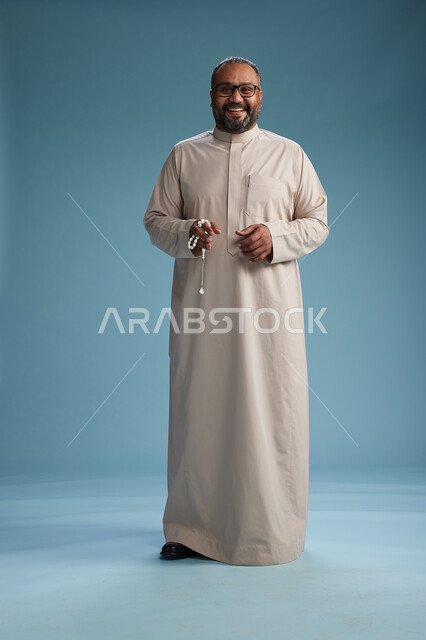 بورتريه لرجل عربي سعودي خليجي مبتسم يرتدي ثوب بيج يمسك بيده سبحه وينظر الى الكاميرا, كامل الجسم, خلفيه زرقاء