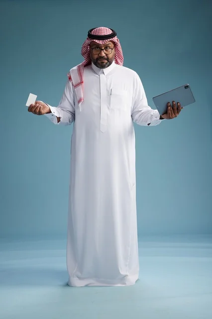 A Saudi man in traditional attire using a tablet and a credit card, the concept of electronic payment, online shopping, blue background, full body.