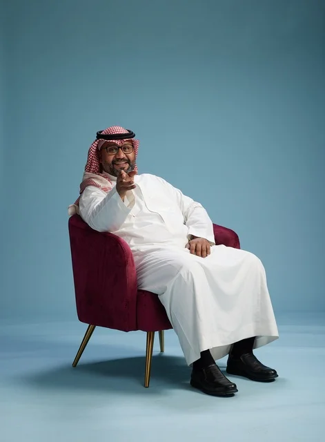 A portrait of a Saudi man wearing a white thobe and a shemagh, gesturing with his hand while sitting on a red chair, with various gestures and movements, against a blue background.