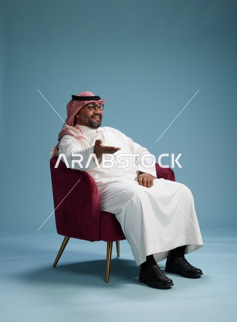 بورتريه لرجل سعودي يرتدي الثوب الابيض والشماغ يُشير بيده أثناء جلوسه على كرسي احمر, ايماءات وحركات منوعه, خلفيه زرقاء