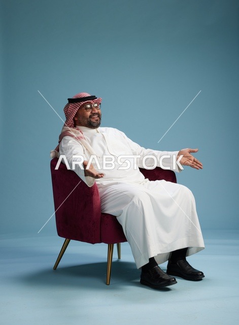 بورتريه لرجل سعودي يرتدي الثوب الابيض والشماغ يُشير بيده أثناء جلوسه على كرسي احمر, ايماءات وحركات منوعه, خلفيه زرقاء