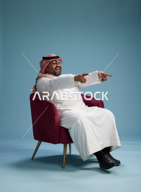 بورتريه لرجل سعودي يرتدي الثوب الابيض والشماغ يُشير بيده أثناء جلوسه على كرسي احمر, ايماءات وحركات منوعه, خلفيه زرقاء
