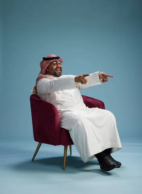 A portrait of a Saudi man wearing a white thobe and a shemagh, gesturing with his hand while sitting on a red chair, with various gestures and movements, against a blue background.
