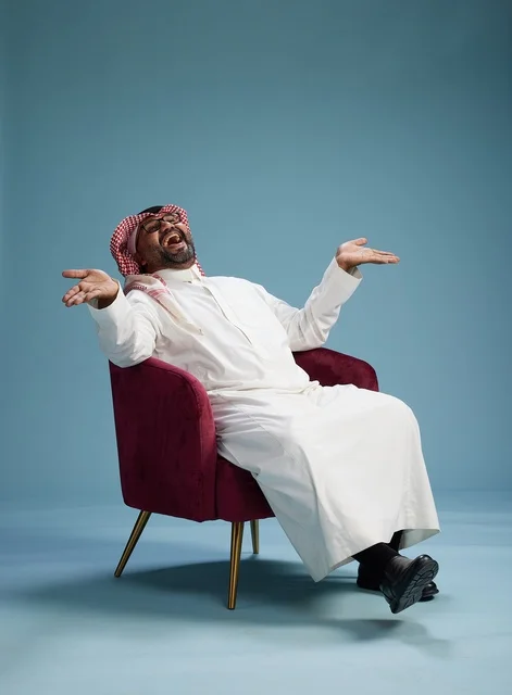 A portrait of a Saudi man wearing a white thobe and a shemagh, gesturing with his hand while sitting on a red chair, with various gestures and movements, against a blue background.