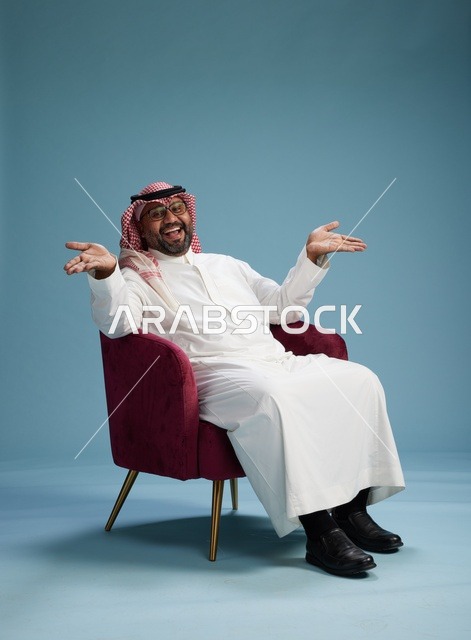 بورتريه لرجل سعودي يرتدي الثوب الابيض والشماغ يُشير بيده أثناء جلوسه على كرسي احمر, ايماءات وحركات منوعه, خلفيه زرقاء