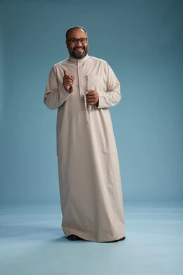 A portrait of a smiling Arab Saudi Gulf man wearing a beige thobe, holding a rosary in his hand and looking at the camera, full body, with a blue background.