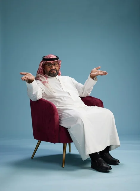 بورتريه لرجل سعودي يرتدي الثوب الابيض والشماغ يُشير بيده أثناء جلوسه على كرسي احمر, ايماءات وحركات منوعه, خلفيه زرقاء