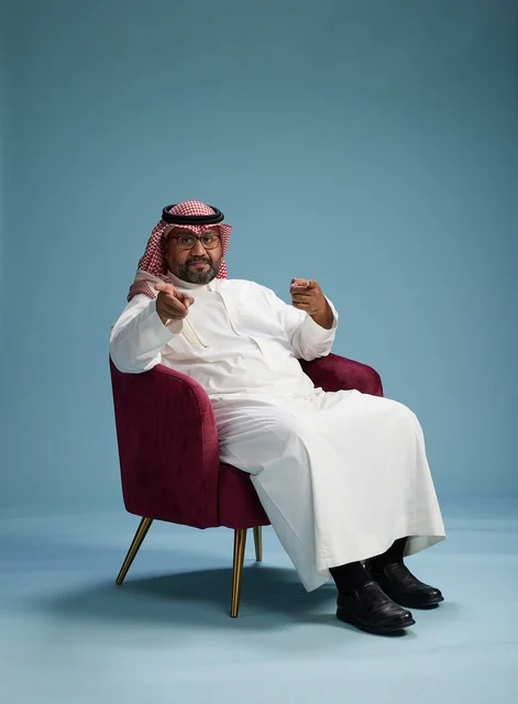 A portrait of a Saudi man wearing a white thobe and a shemagh, gesturing with his hand while sitting on a red chair, with various gestures and movements, against a blue background.