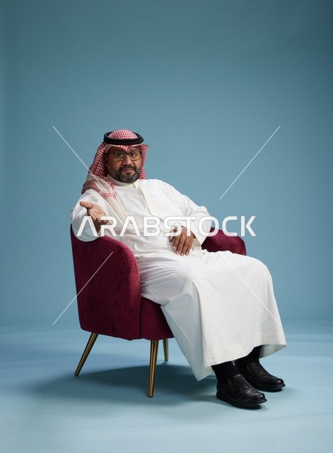 بورتريه لرجل سعودي يرتدي الثوب الابيض والشماغ يُشير بيده أثناء جلوسه على كرسي احمر, ايماءات وحركات منوعه, خلفيه زرقاء