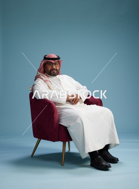 بورتريه لرجل سعودي يرتدي الثوب الابيض والشماغ يُشير بيده أثناء جلوسه على كرسي احمر, ايماءات وحركات منوعه, خلفيه زرقاء
