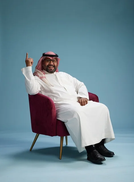 A portrait of a Saudi man wearing a white thobe and a shemagh, gesturing with his hand while sitting on a red chair, with various gestures and movements, against a blue background.