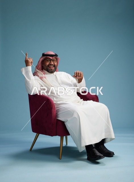 A portrait of a Saudi man wearing a white thobe and a shemagh, gesturing with his hand while sitting on a red chair, with various gestures and movements, against a blue background.
