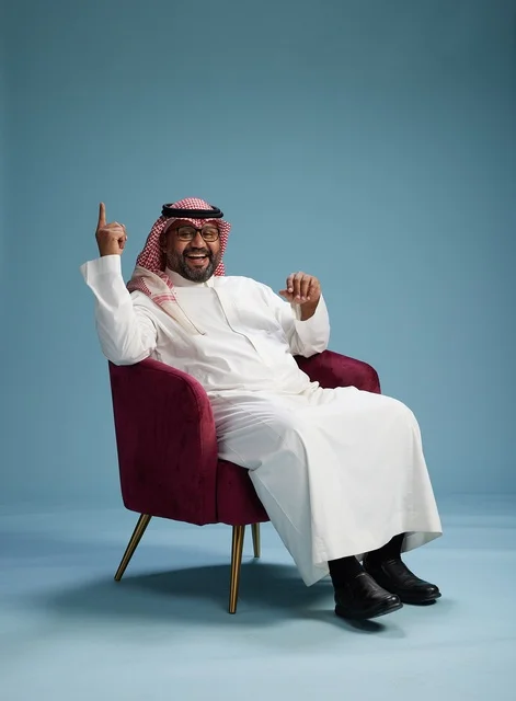 A portrait of a Saudi man wearing a white thobe and a shemagh, gesturing with his hand while sitting on a red chair, with various gestures and movements, against a blue background.