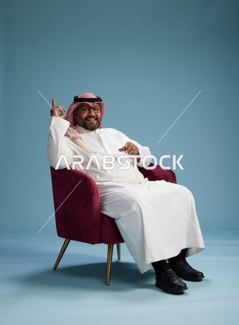 بورتريه لرجل سعودي يرتدي الثوب الابيض والشماغ يُشير بيده أثناء جلوسه على كرسي احمر, ايماءات وحركات منوعه, خلفيه زرقاء