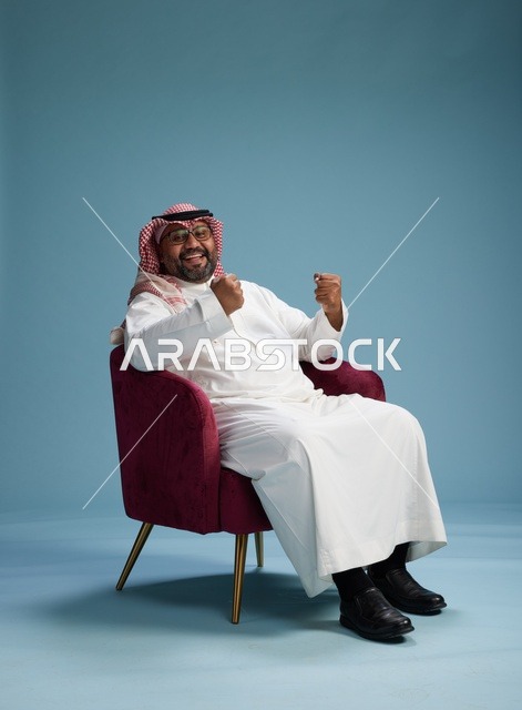 بورتريه لرجل سعودي يرتدي الثوب الابيض والشماغ يُشير بيده أثناء جلوسه على كرسي احمر, ايماءات وحركات منوعه, خلفيه زرقاء