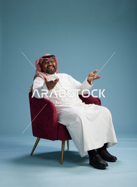 بورتريه لرجل سعودي يرتدي الثوب الابيض والشماغ يُشير بيده أثناء جلوسه على كرسي احمر, ايماءات وحركات منوعه, خلفيه زرقاء