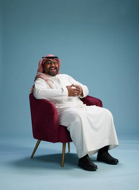 A portrait of a Saudi man wearing a white thobe and a shemagh, gesturing with his hand while sitting on a red chair, with various gestures and movements, against a blue background.