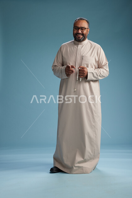 بورتريه لرجل عربي سعودي خليجي مبتسم يرتدي ثوب بيج يمسك بيده سبحه وينظر الى الكاميرا, كامل الجسم, خلفيه زرقاء