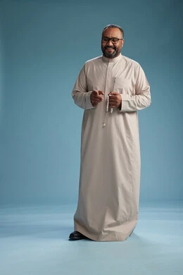 A portrait of a smiling Arab Saudi Gulf man wearing a beige thobe, holding a rosary in his hand and looking at the camera, full body, with a blue background.