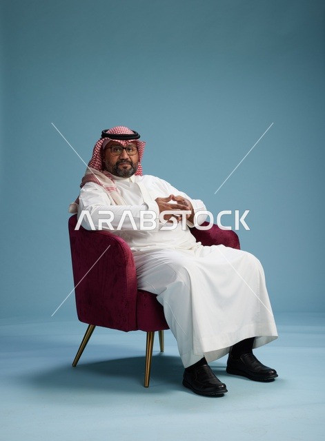 بورتريه لرجل سعودي يرتدي الثوب الابيض والشماغ يُشير بيده أثناء جلوسه على كرسي احمر, ايماءات وحركات منوعه, خلفيه زرقاء