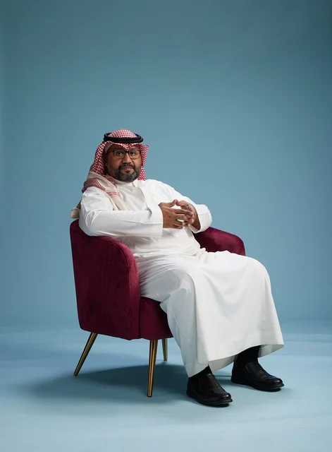 A portrait of a Saudi man wearing a white thobe and a shemagh, gesturing with his hand while sitting on a red chair, with various gestures and movements, against a blue background.