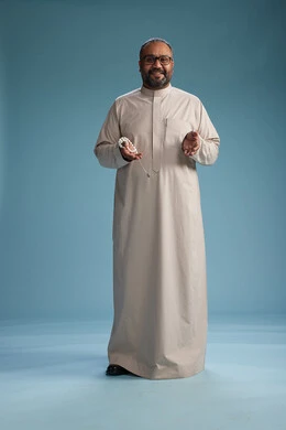 A portrait of a smiling Arab Saudi Gulf man wearing a beige thobe, holding a rosary in his hand and looking at the camera, full body, with a blue background.