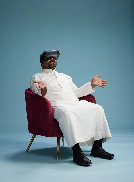 A Saudi man in traditional attire is experiencing virtual reality, watching VR movies, with a blue background.