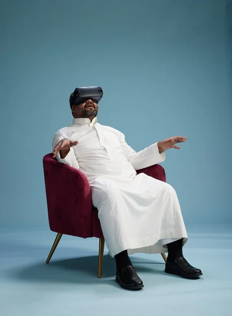 A Saudi man in traditional attire is experiencing virtual reality, watching VR movies, with a blue background.