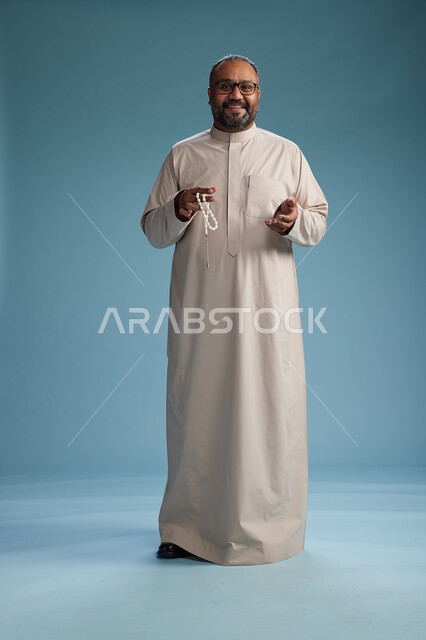 بورتريه لرجل عربي سعودي خليجي مبتسم يرتدي ثوب بيج يمسك بيده سبحه وينظر الى الكاميرا, كامل الجسم, خلفيه زرقاء