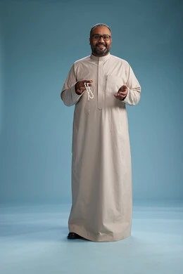 بورتريه لرجل عربي سعودي خليجي مبتسم يرتدي ثوب بيج يمسك بيده سبحه وينظر الى الكاميرا, كامل الجسم, خلفيه زرقاء